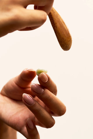 Close-up of a hand holding a small quantity of Peach and Cream vulva balm with another hand holding a wooden stick against a beige background.