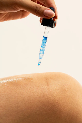 Hand holding a dropper with Peach and Cream 'Après' after shave blue liquid above skin