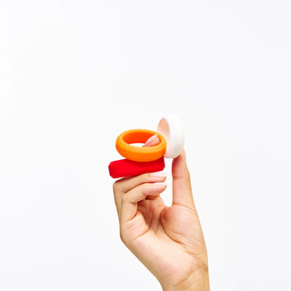 Hand holding Peach and Cream 'O' intimate silicone ring set, one red and one orange, one white, on a white background.