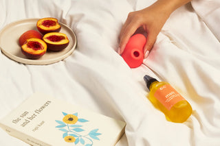 Hand holding Peach and Cream 'aura' vibrator next to a bottle of massage oil 'melt' on bed sheets with peaches and a book.