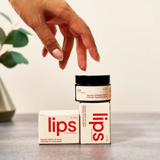 Hand reaching towards a jar labeled Peach and Cream vulva balm 'lips' on a neutral background